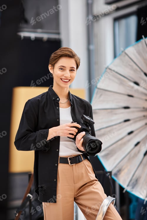 Preview: happy beautiful woman in casual attire smiling at camera and holding camera in her photo studio
