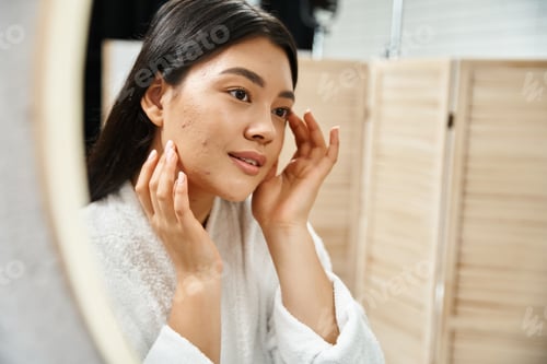 Preview: young asian woman with brunette hair examining her skin in the bathroom mirror, skin condition