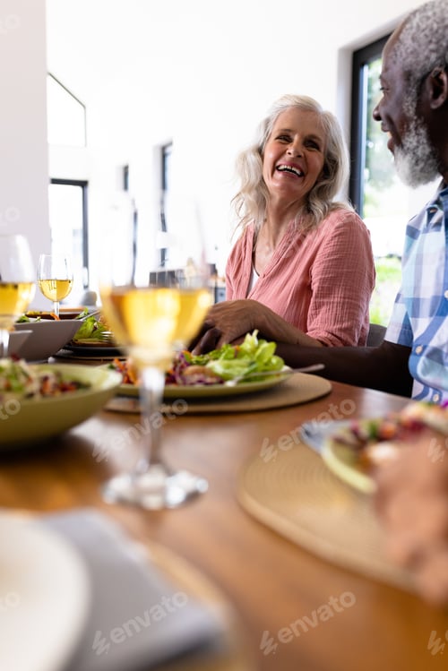 Preview: Cheerful multiracial senior couple holding hands while having lunch at dining table in nursing home