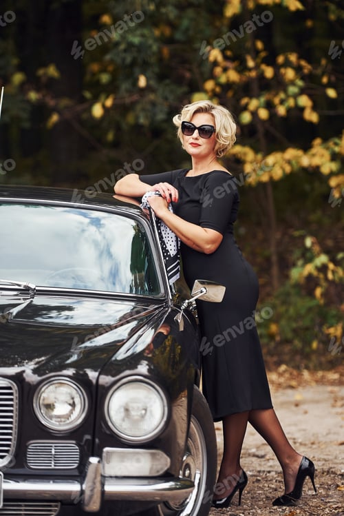 Preview: Blonde woman in sunglasses and in black dress near old vintage classic car