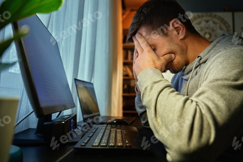 Preview: Exhausted man at home office with hands on face after long work hours
