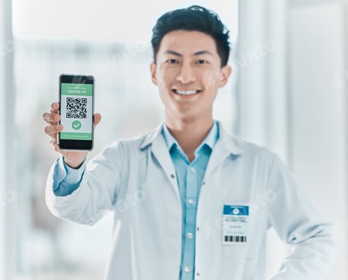Preview: Shot of a young doctor standing alone in the clinic and showing his Covid passport on his cellphone