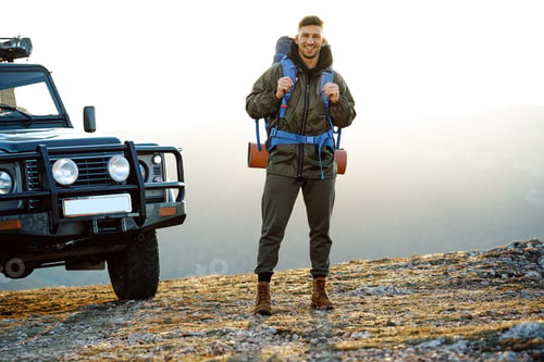 Preview: Portrait of a young traveler man in hiking equipment standing near his off-road car