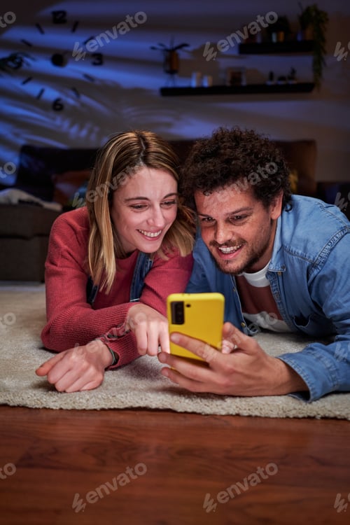 Preview: Young couple lying down on the living room floor using phone together, watching social media app.