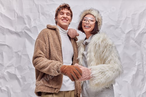 Preview: trendy multiethnic couple in stylish winter clothes smiling at camera on white textured backdrop