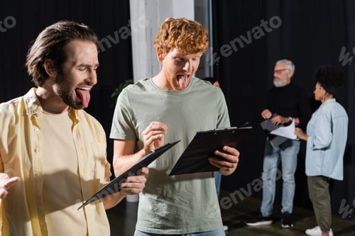Preview: brunette and redhead men sticking out tongues while reading scenarios during rehearsal in theater
