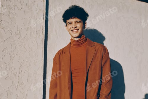 Preview: Smiling young man in a cozy sweater and blazer against a light textured wall The warm colors create