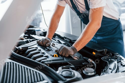 Preview: Quality service. Young man in white shirt and blue uniform repairs automobile