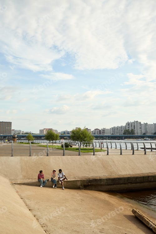 Preview: Group of Multiethnic Teenagers Sitting Outdoors near Urban Waterfront Talking