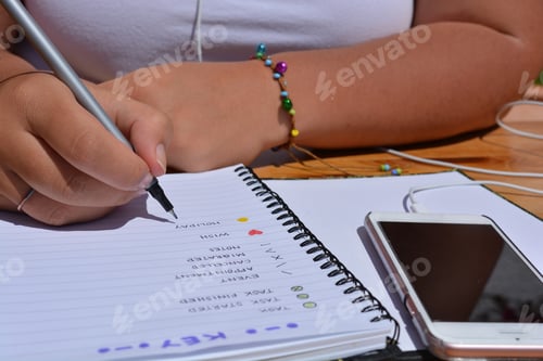 Preview: millennial woman writing in bullet journal with a smartphone on the desk beside her