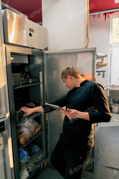 Preview: chef stands in kitchen in a black uniform and looks through what is on the list in the refrigerator
