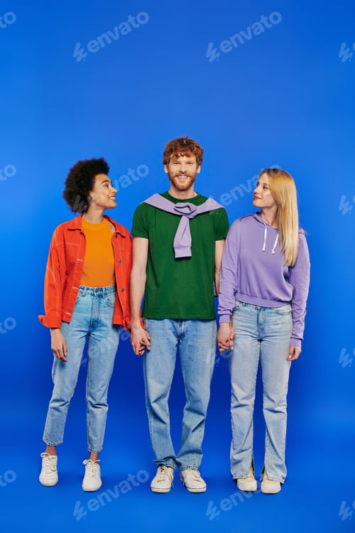 Preview: polyamory three people, young man and women holding hands on blue background, studio shot, vibrant
