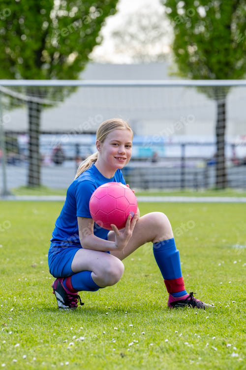 Preview: Young female soccer player in a blue uniform kneeling on grass field with a pink soccer ball.