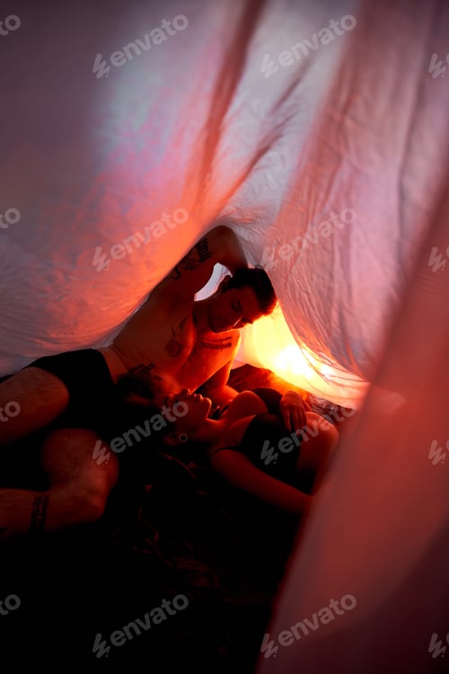 Preview: sexy multicultural couple in underwear lying sensually together under bedsheet surrounded by lights