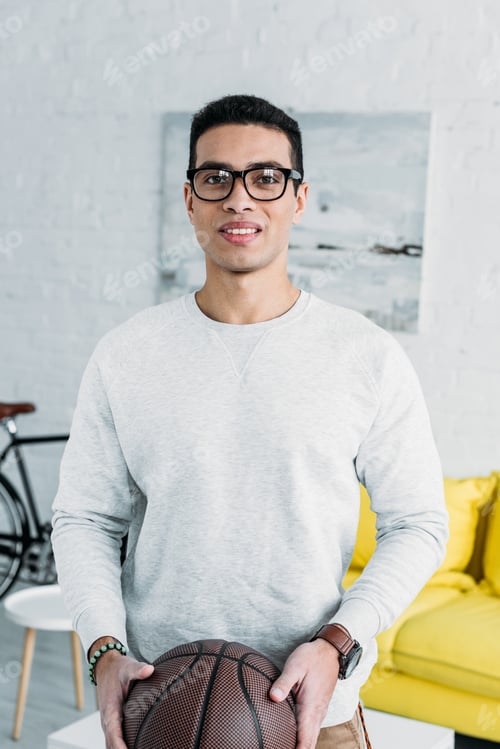 Preview: handsome mixed race man in white sweatshirt and glasses holding brown ball