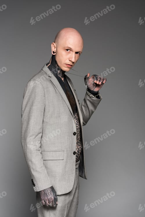 Preview: side view of handsome bald tattooed man in suit holding dog tag on chain and looking at camera