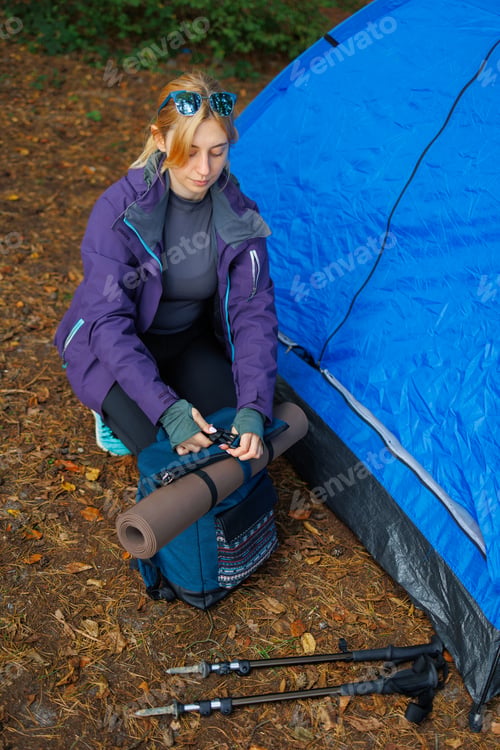 Preview: Woman in the forest near a tent attaches a yoga mat to a backpack before going on a hike