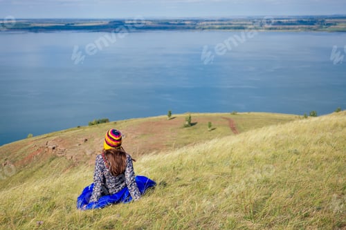 Preview: happy girl in a funny hat from Nepal sitting in a blue sleeping bag and looks at the lake landscape