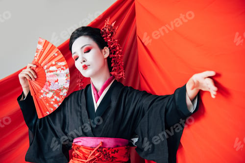 Preview: beautiful geisha in black kimono with hand fan and red cloth on background dancing isolated on white