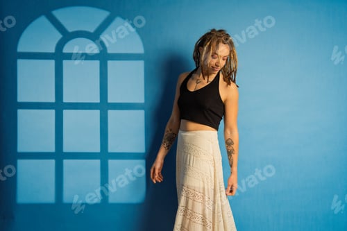 Preview: Woman with dreadlocks and tattoos posing against blue wall