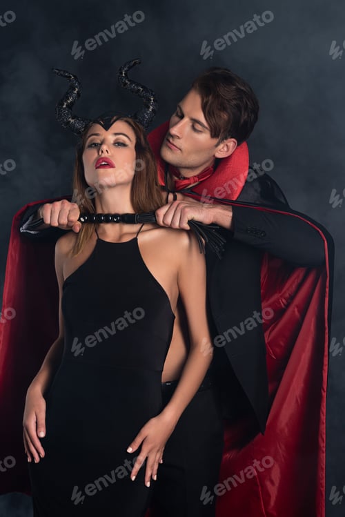 Preview: handsome man in vampire halloween costume holding flogging whip near girl with horns on black with