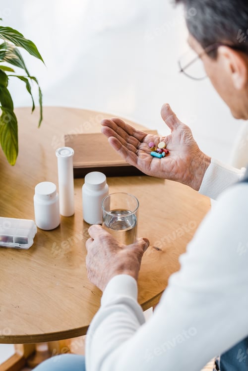 Preview: selective focus of retired man looking at pills while holding glass of water