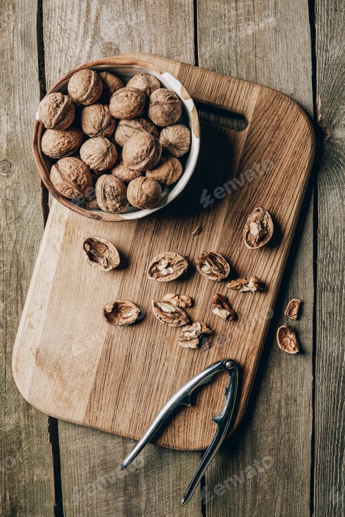Preview: top view of walnuts in bowl and nutcracker on chopping board on wooden table