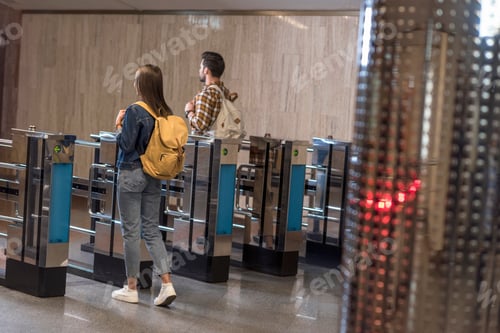 Preview: rear view of couple of stylish tourists with backpacks passing through turnstiles
