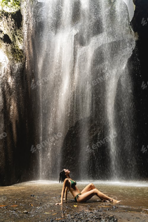 Preview: Pleased brunette girl sitting under the waterfall