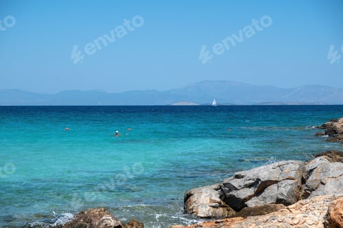 Preview: Greece Agistri Island. Rocky beach, people swimming sailboat sails in sea water, blue sky sunny day