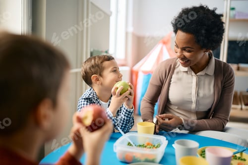 Preview: Happy black teacher and small boy talking during lunch time at kindergarten.