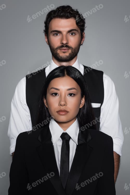 Preview: young asian woman and brunette man in formal wear looking at camera isolated on grey