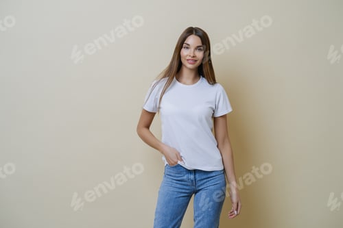 Preview: Studio shot of pretty slim woman keeps hand in pocket, wears casual t shirt and jeans