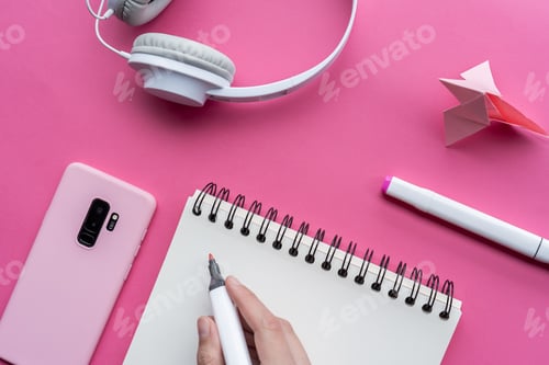 Preview: High angle shot of a cellphone, headphones, and writing tools on a pink surface