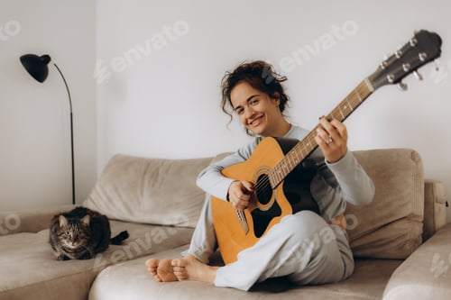 Preview: Brunette attractive female smiling and learning to play guitar while sitting in cozy home interior