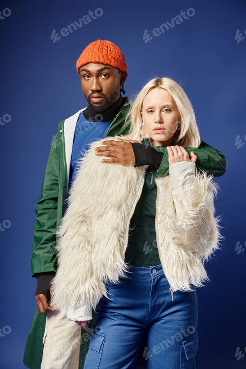 Preview: african american man embracing blonde woman in stylish winter attire on blue backdrop, multiethnic
