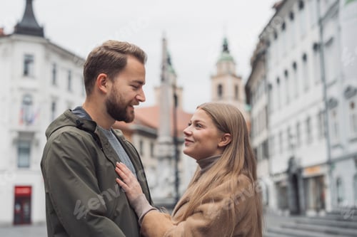 Preview: A couple stands close together in a charming European square, smiling at each other. The woman has l