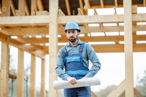 Preview: Builder with blueprints near the wooden house on the construction site