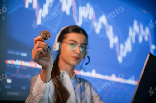 Preview: Holding bitcoin in hand. Female stock broker is working indoors. Big wide display behind her