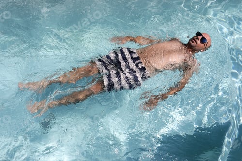 Preview: Young bald handsome man floating in a swimming pool wearing sunglasses
