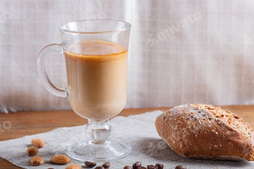 Preview: Glass cup of coffee with cream and bun on a wooden background and linen textile.