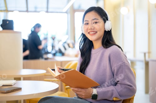 Preview: Asian woman listening to music in headphones and reading a book while sitting at round table in cafe