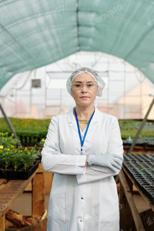 Preview: Serious mature female scientist in workwear standing in large hothouse