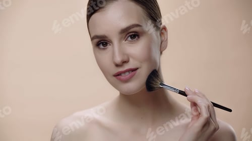 Preview: joyful woman with bare shoulders applying face powder while holding cosmetic brush isolated on beige