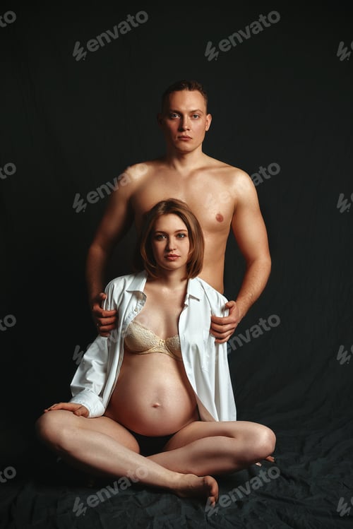 Preview: A pregnant couple posing at the camera on a dark background, low key. Fashion Pregnancy Concept