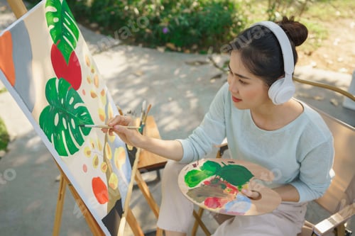 Preview: Outdoor activity concept, Female artist listening music while painting picture on canvas in garden
