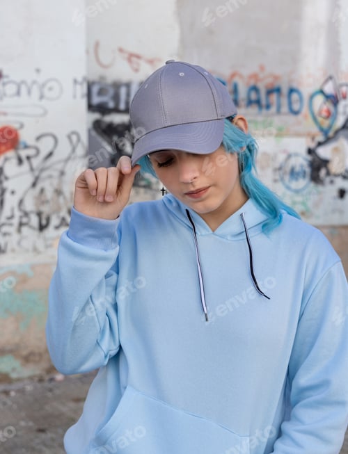 Preview: Blue haired Teenage girl in blue hoodie and baseball cap staying against graffiti wall