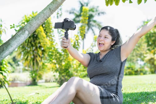 Preview: young latin influencer woman sitting on the grass of the park recording a video with great happiness