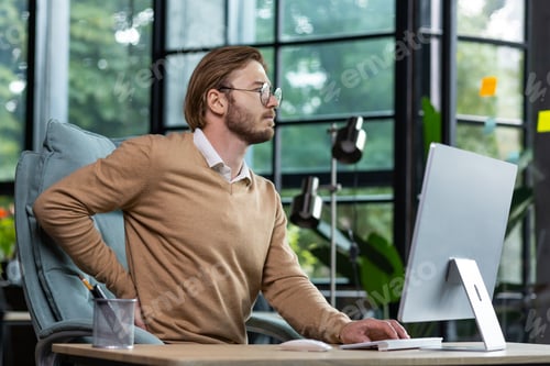 Preview: A young man is tired at the workplace in the office, massages his back with his hand, overtime work