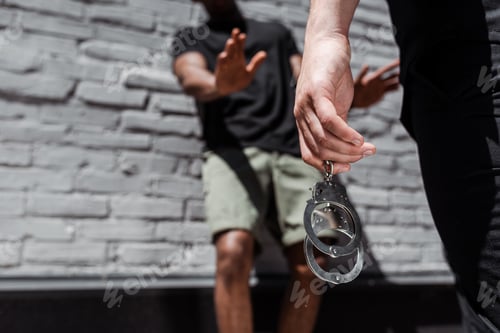 Preview: cropped view of police officer holding handcuffs while detaining african american man gesturing near
